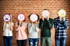 5 people stood in front of a brick wall. You can't see their faces as they are all holding up an emoji style face mask in front of them. The emotions range from angry (on the left) up to delighted (on the right).